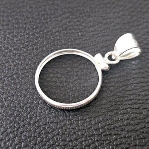 May include: A silver-colored pendant with a circular frame and a loop for a chain. The pendant has a small decorative element where the loop connects to the frame. The background is a textured black surface.