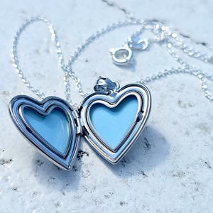 May include: A silver heart-shaped locket necklace with a delicate chain. The locket is open, revealing a light blue interior. The necklace is displayed on a textured, light-colored surface.