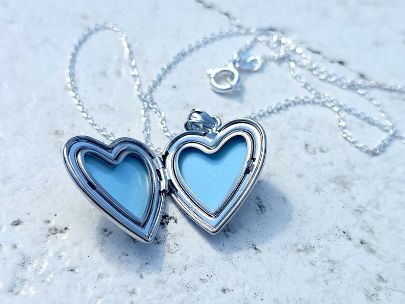 May include: A silver heart-shaped locket necklace with a delicate chain. The locket is open, revealing a light blue interior. The necklace is displayed on a textured, light-colored surface.