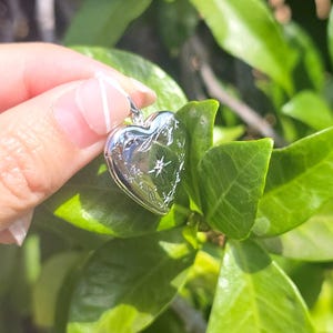 May include: A silver heart-shaped locket with engraved floral and starburst designs. The locket is held against a backdrop of green leaves, suggesting a nature-inspired theme. The locket's surface reflects light, highlighting its polished finish.