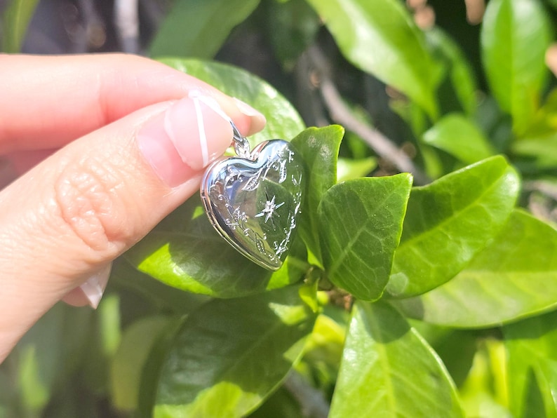May include: A silver heart-shaped locket with engraved floral and starburst designs. The locket is held against a backdrop of green leaves, suggesting a nature-inspired theme. The locket's surface reflects light, highlighting its polished finish.