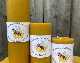 May include: Three golden yellow beeswax candles of varying heights. Each candle has a white label with the text "Greenbrier Golden Honey Handmade Beeswax Candle" and a graphic of a bee on a honeycomb. The candles are cylindrical.