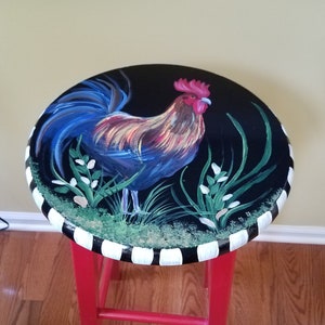 May include: A red painted stool with a black and white striped top. The top features a hand-painted rooster with a blue tail and red comb, standing in a field of green grass with white flowers.