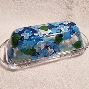 Può includere: Un porta burro in vetro trasparente con un design dipinto a mano di fiori blu e bianchi. I fiori sono dipinti in uno stile realistico e sono circondati da foglie verdi.