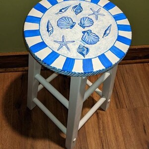 Op de afbeelding: Witte kruk met een blauw-wit gestreept bovenblad. Het bovenblad is beschilderd met schelpen en zeesterren.