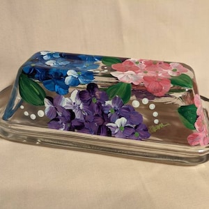 Puede incluir: Un recipiente de mantequilla de vidrio con un diseño pintado a mano de flores moradas, rosas y azules. El recipiente tiene un fondo blanco con puntos blancos.