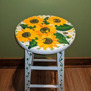 May include: White wooden stool with a painted design of sunflowers and green leaves. The stool has a white base and green vines painted on the legs.