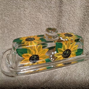 May include: A clear glass butter dish with a lid, hand-painted with yellow sunflowers and green leaves. The sunflowers have dark brown centres.