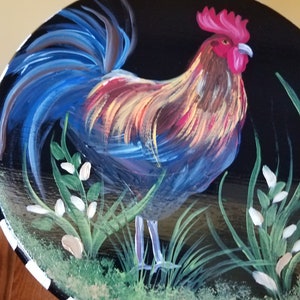 May include: Hand-painted wooden table top with a rooster standing in a field of green grass and white flowers. The rooster has a red comb and wattle, blue and yellow feathers, and a long blue tail.