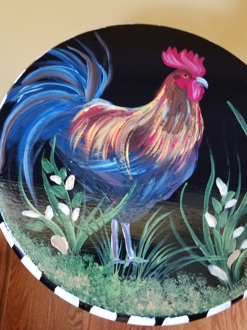 May include: Hand-painted wooden table top with a rooster standing in a field of green grass and white flowers. The rooster has a red comb and wattle, blue and yellow feathers, and a long blue tail.
