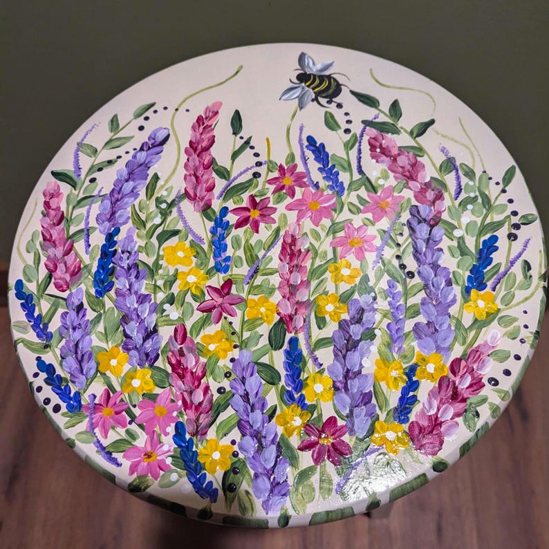 May include: A hand-painted wooden stool with a floral design. The stool features a variety of flowers in shades of pink, purple, yellow, and green, with a bee painted in the center.