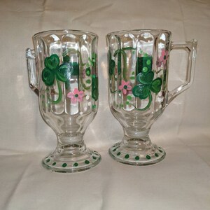 May include: Two clear glass mugs with green shamrock and pink flower designs. The mugs have a fluted design and a green polka dot trim.