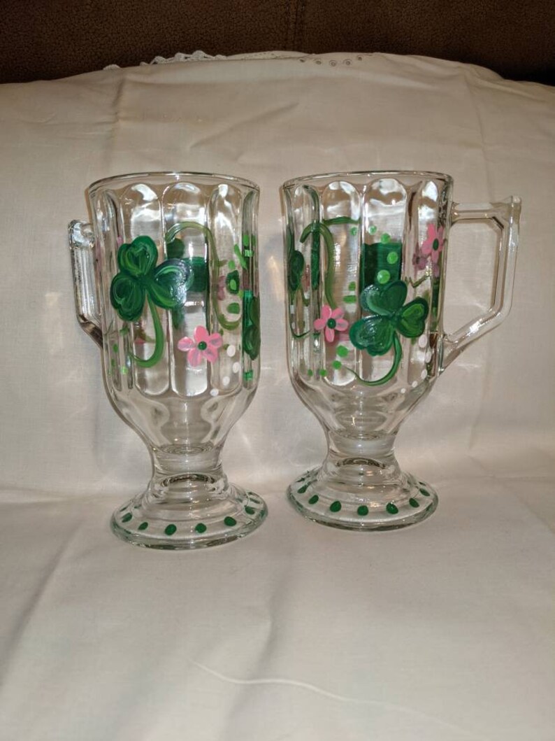 May include: Two clear glass mugs with green shamrock and pink flower designs. The mugs have a fluted design and a green polka dot trim.