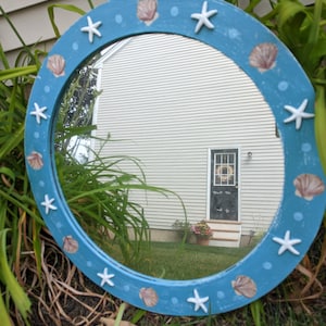 May include: A round blue mirror with a white painted seashell and starfish design. The mirror is reflecting a house with a door and a wreath.