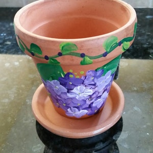 Pode incluir: Um vaso de flores de terracota com um desenho pintado de flores roxas e folhas verdes. O vaso tem um pires correspondente.