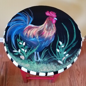 May include: A red painted stool with a black and white striped base. The top of the stool is painted black with a rooster design in shades of blue, red, yellow, and green.