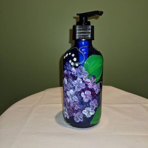 Könnte beinhalten: Ein blauer Glas-Seifenspender mit schwarzem Pumpkopf. Die Flasche ist handbemalt mit lila und weißen Blumen und grünen Blättern.
