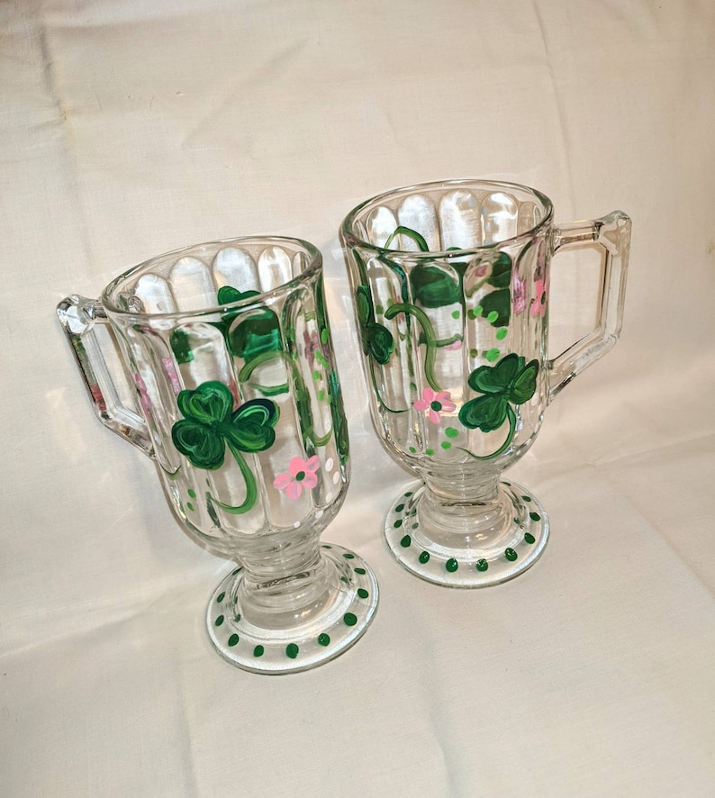 May include: Two clear glass mugs with a footed base. The mugs are decorated with hand-painted green shamrocks and pink flowers.