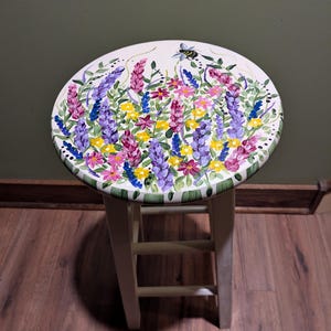 May include: White wooden stool with a round seat painted with a floral design. The design features purple, pink, yellow, and blue flowers with green leaves and a bee.