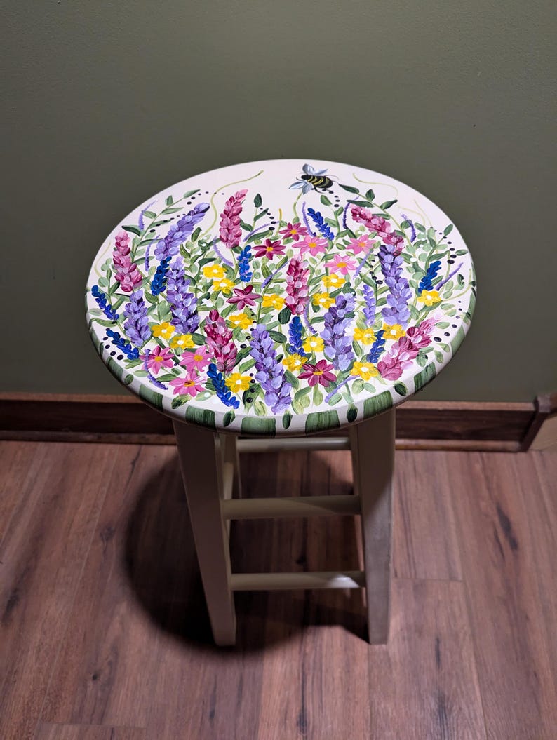 May include: White wooden stool with a round seat painted with a floral design. The design features purple, pink, yellow, and blue flowers with green leaves and a bee.
