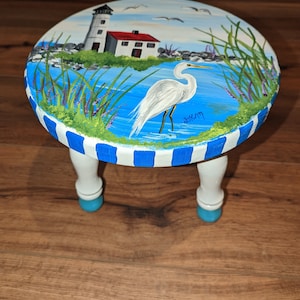 Puede incluir: Un taburete de madera pintado de blanco con rayas azules y blancas alrededor de la parte superior. La parte superior del taburete está pintada con una escena de un faro, una garceta blanca y una masa de agua. El taburete tiene tres patas con pies pintados de azul.