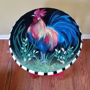 May include: A hand-painted wooden stool with a black and white striped base. The top of the stool features a colorful rooster painted on a black background.