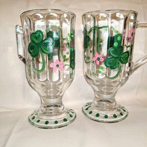 May include: Two clear glass mugs with green shamrock and pink flower designs. The mugs have a textured, fluted design and a green polka dot base.