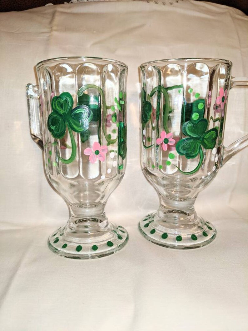 May include: Two clear glass mugs with green shamrock and pink flower designs. The mugs have a textured, fluted design and a green polka dot base.