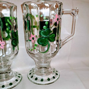 May include: Two clear glass mugs with a footed base. The mugs are decorated with hand-painted green shamrocks, pink flowers, and white dots.