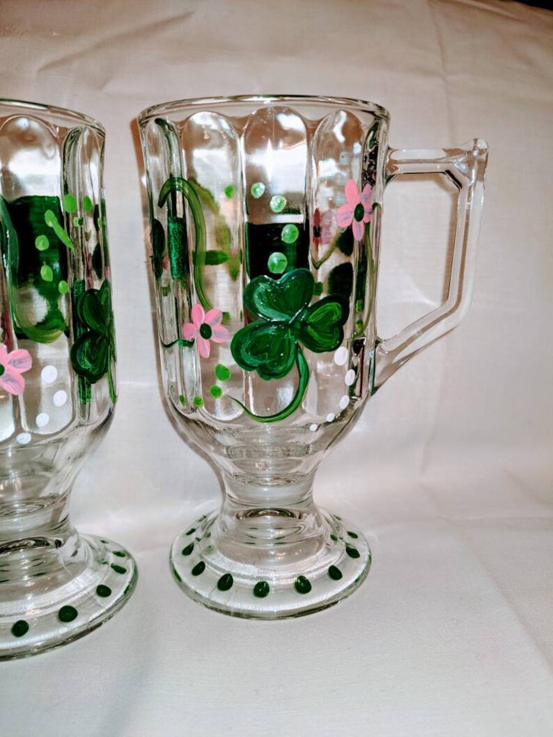 May include: Two clear glass mugs with a footed base. The mugs are decorated with hand-painted green shamrocks, pink flowers, and white dots.