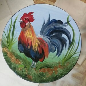 K&ouml;nnte beinhalten: Eine handbemalte Holztischplatte mit einem Hahn-Design. Der Hahn ist in Rot-, Gelb-, Blau- und Schwarzt&ouml;nen bemalt. Der Hintergrund ist hellblau mit gr&uuml;nem Gras und braunem Boden.