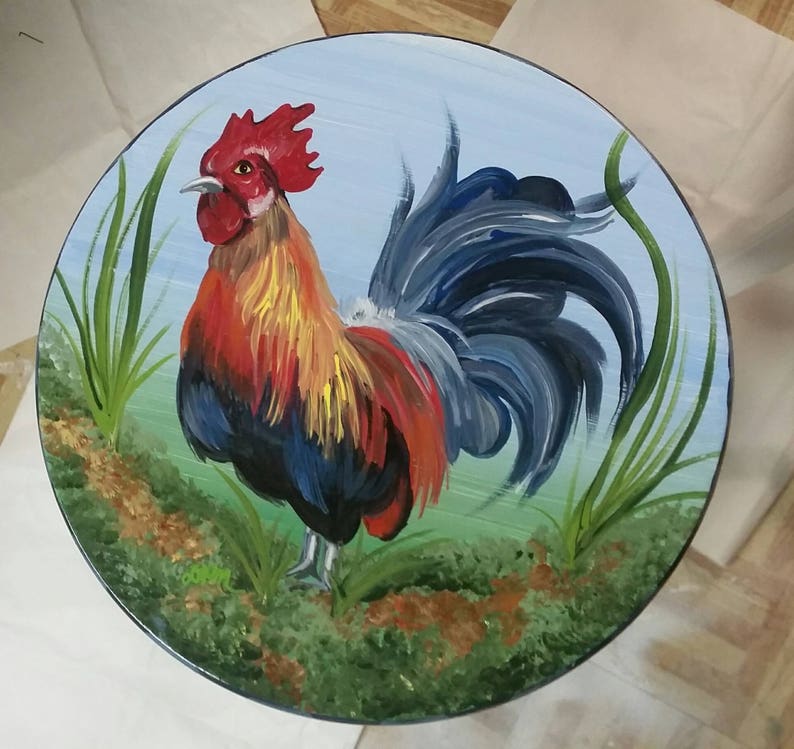 K&ouml;nnte beinhalten: Eine handbemalte Holztischplatte mit einem Hahn-Design. Der Hahn ist in Rot-, Gelb-, Blau- und Schwarzt&ouml;nen bemalt. Der Hintergrund ist hellblau mit gr&uuml;nem Gras und braunem Boden.