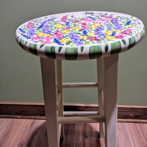 May include: A white wooden stool with a round seat painted with a floral design. The design features pink, purple, yellow, and green flowers.