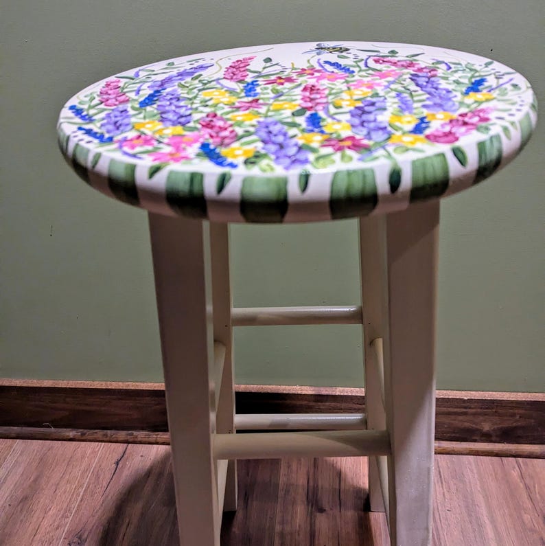 May include: A white wooden stool with a round seat painted with a floral design. The design features pink, purple, yellow, and green flowers.