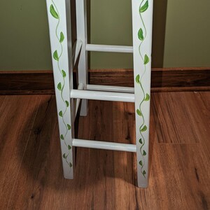 May include: White wooden stool with green leaf vines painted on the side. The stool has three rungs.
