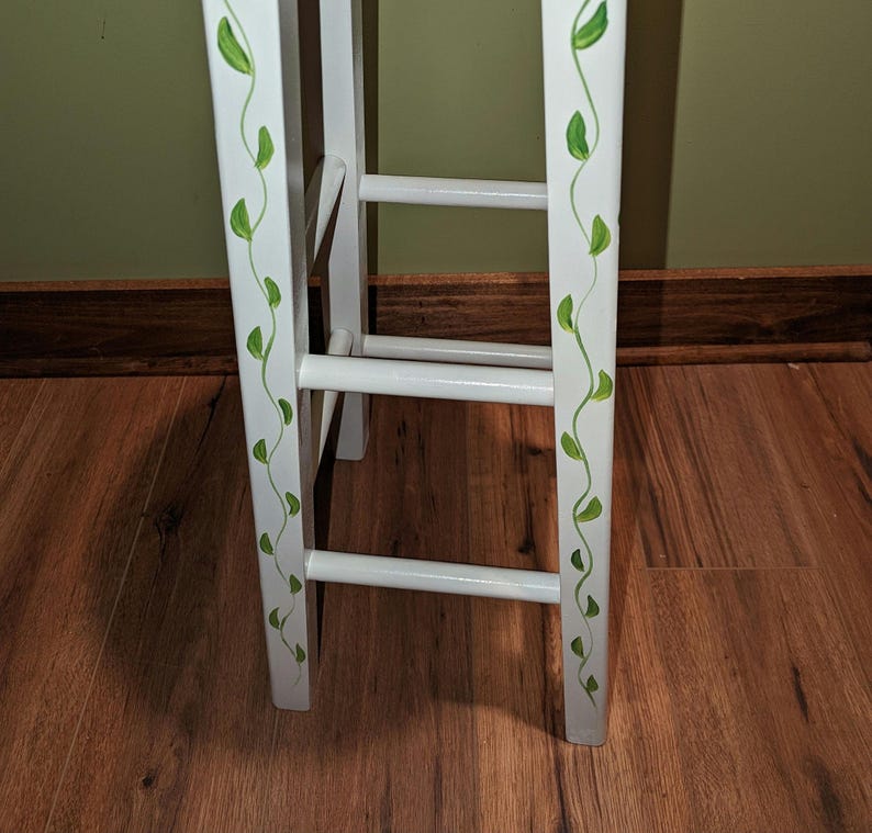 May include: White wooden stool with green leaf vines painted on the side. The stool has three rungs.