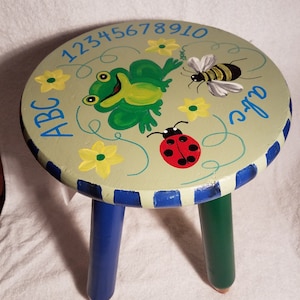 Puede incluir: Un taburete de madera pintado a mano con tres patas. La parte superior del taburete está pintada de verde con una rana, una mariquita, una abeja y flores amarillas. Los números 12345678910 están pintados alrededor del borde del taburete. Las letras ABC están pintadas en el lado izquierdo del taburete. Las palabras "Little" y "One" están pintadas en el lado derecho del taburete.
