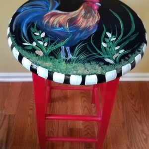 May include: A red painted stool with a black and white striped top. The top features a hand-painted rooster with blue, red, yellow, and green accents.
