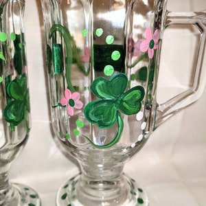 May include: Two clear glass mugs with green shamrock and pink flower designs painted on them. The mugs have a white rim and a green and white polka dot base.