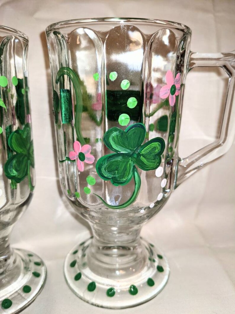 May include: Two clear glass mugs with green shamrock and pink flower designs painted on them. The mugs have a white rim and a green and white polka dot base.