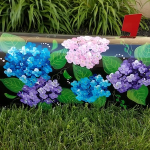 May include: A black mailbox painted with a floral design featuring hydrangeas in shades of blue, purple, and pink. The flowers are surrounded by green leaves.