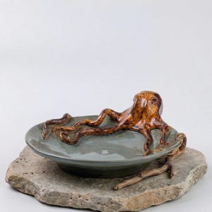 以下が含まれることがあります： 茶色の釉薬を施したセラミック製のタコの彫刻が、グレーグリーンの皿の上に置かれています。 皿は、質感のある灰色の石の台座の上にあります。 タコは詳細な触手を持っています。