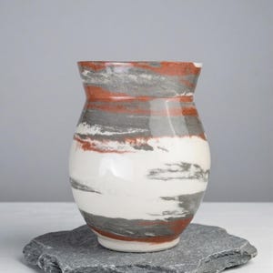 May include: A ceramic vase with a wide, rounded body and a slightly flared rim. The vase features horizontal bands of white, gray, and reddish-brown colors. It sits on a gray, irregularly shaped stone.