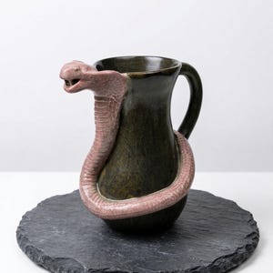 May include: A dark green ceramic pitcher with a handle, featuring a pink cobra wrapped around the body. The cobra's head forms the spout. The pitcher sits on a round, dark gray slate coaster.