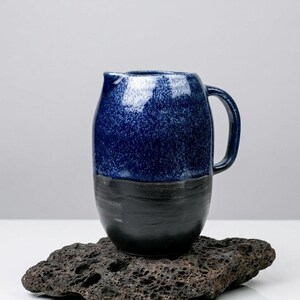 May include: A handcrafted ceramic pitcher with a dark blue speckled upper half and a solid black lower half. The pitcher has a curved handle and a spout for pouring. It sits on a dark, textured stone.