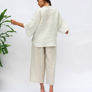 May include: A white linen shirt with thin black stripes and a relaxed fit. The shirt has a collar and three-quarter sleeves. The model is wearing the shirt with beige linen pants.