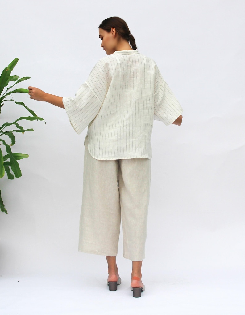 May include: A white linen shirt with thin black stripes and a relaxed fit. The shirt has a collar and three-quarter sleeves. The model is wearing the shirt with beige linen pants.