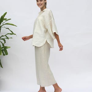 May include: A white linen shirt with thin black stripes and a relaxed fit. The shirt has a collar, a button closure, and a patch pocket on the left side. The model is wearing the shirt with white linen pants.