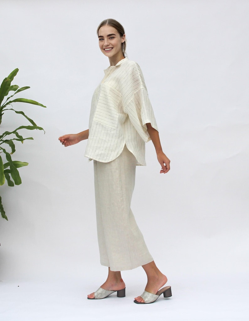May include: A white linen shirt with thin black stripes and a relaxed fit. The shirt has a collar, a button closure, and a patch pocket on the left side. The model is wearing the shirt with white linen pants.