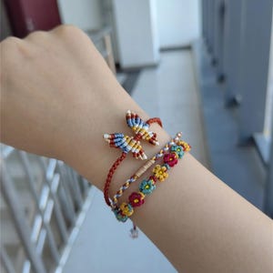 Könnte beinhalten: Drei bunte Freundschaftsarmbänder mit einem Schmetterlingsanhänger. Die Armbänder sind aus roter, blauer, gelber und weißer Schnur gefertigt. Der Schmetterlingsanhänger ist aus mehrfarbiger Schnur gefertigt und hat ein rot-weiß-blaues Muster.